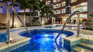 A swimming pool with metal railings, surrounded by palm trees and a multi-story building with balconies.