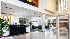 A modern hotel lobby with a reception desk labeled "MONACO," a seating area, and a large window offering a view of the outdoors.