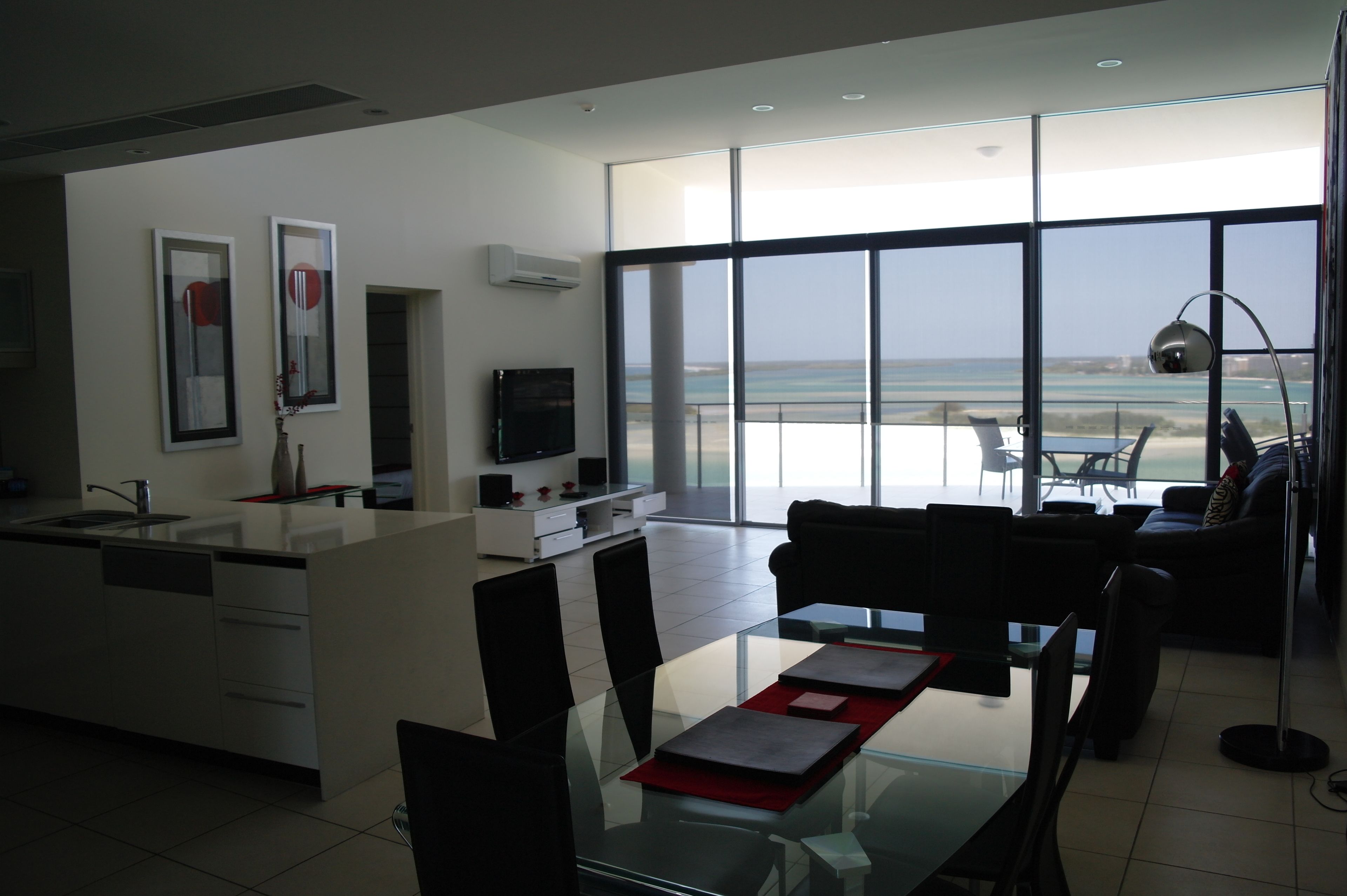 Ein modernes Wohnzimmer mit einem Glastisch, einer Couch und einem Fernseher. Ein großes Fenster bietet Blick auf den Strand.