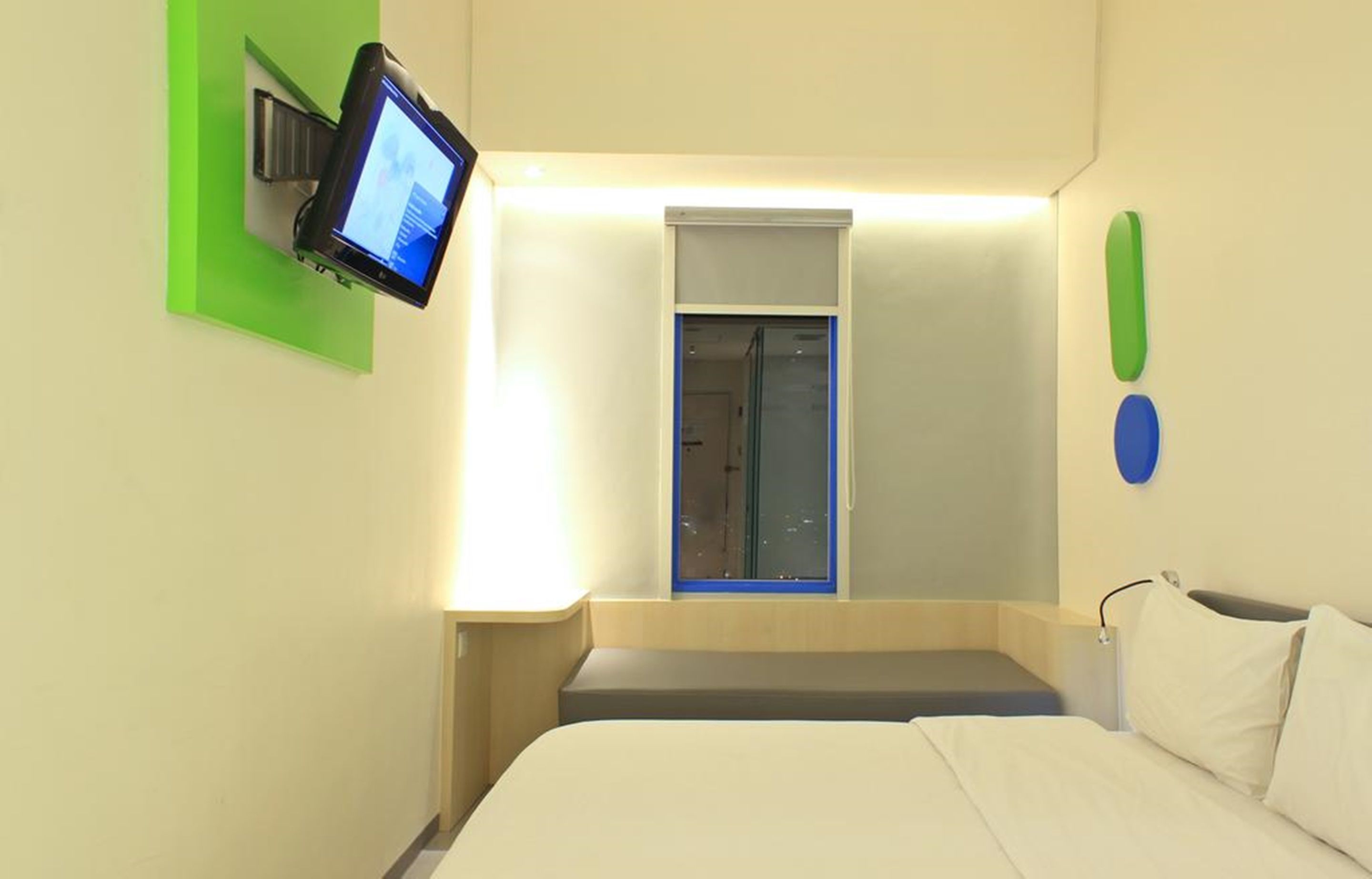A hotel room with a bed, a mounted TV, a small window, and a wall-mounted light fixture.