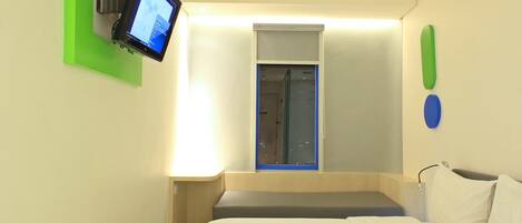 A hotel room with a bed, a mounted TV, a small window, and a wall-mounted light fixture.