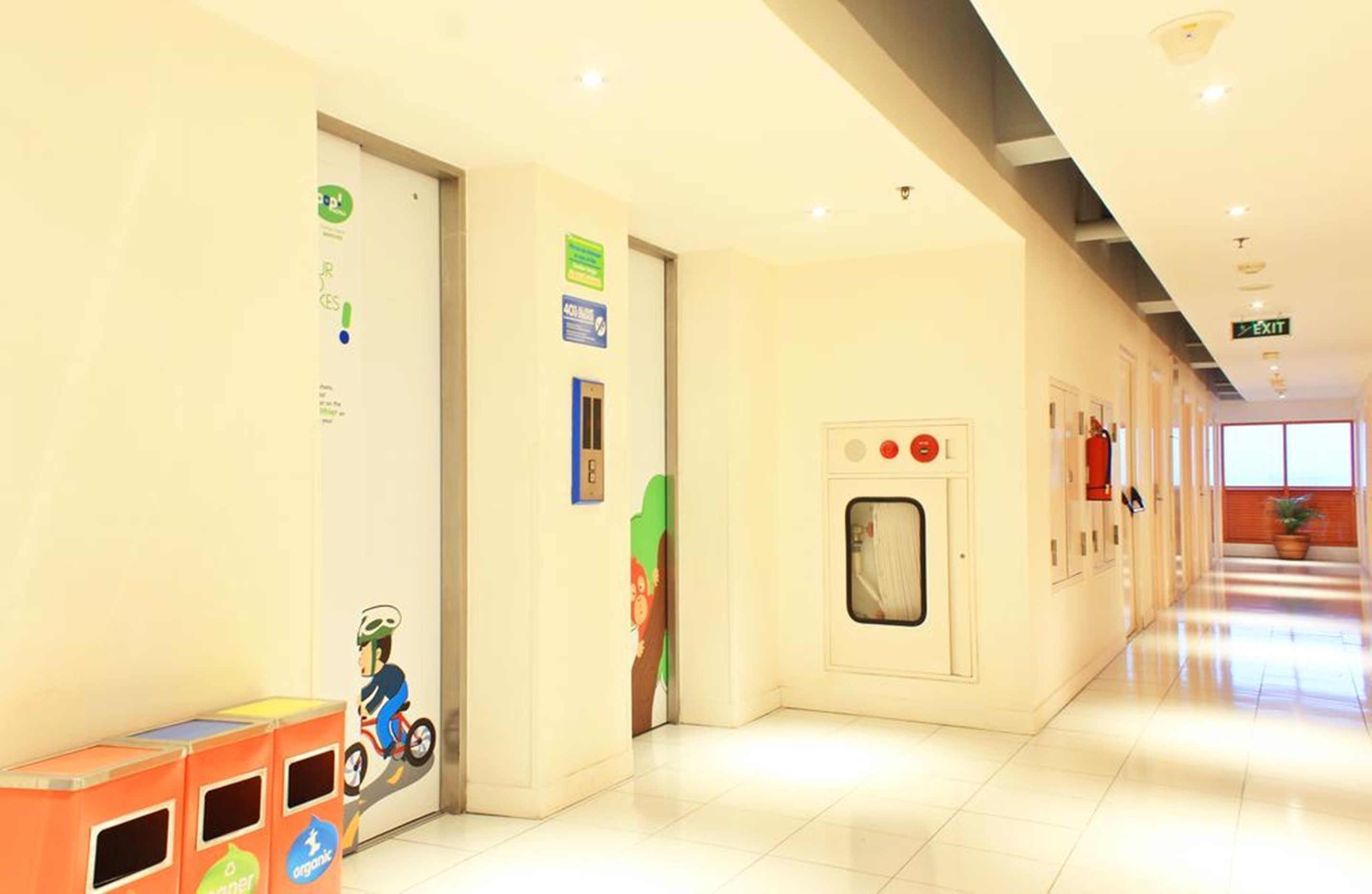 Corredor limpo e bem iluminado, com um contentor de lixo, extintor de incêndio e uma porta com placa verde.