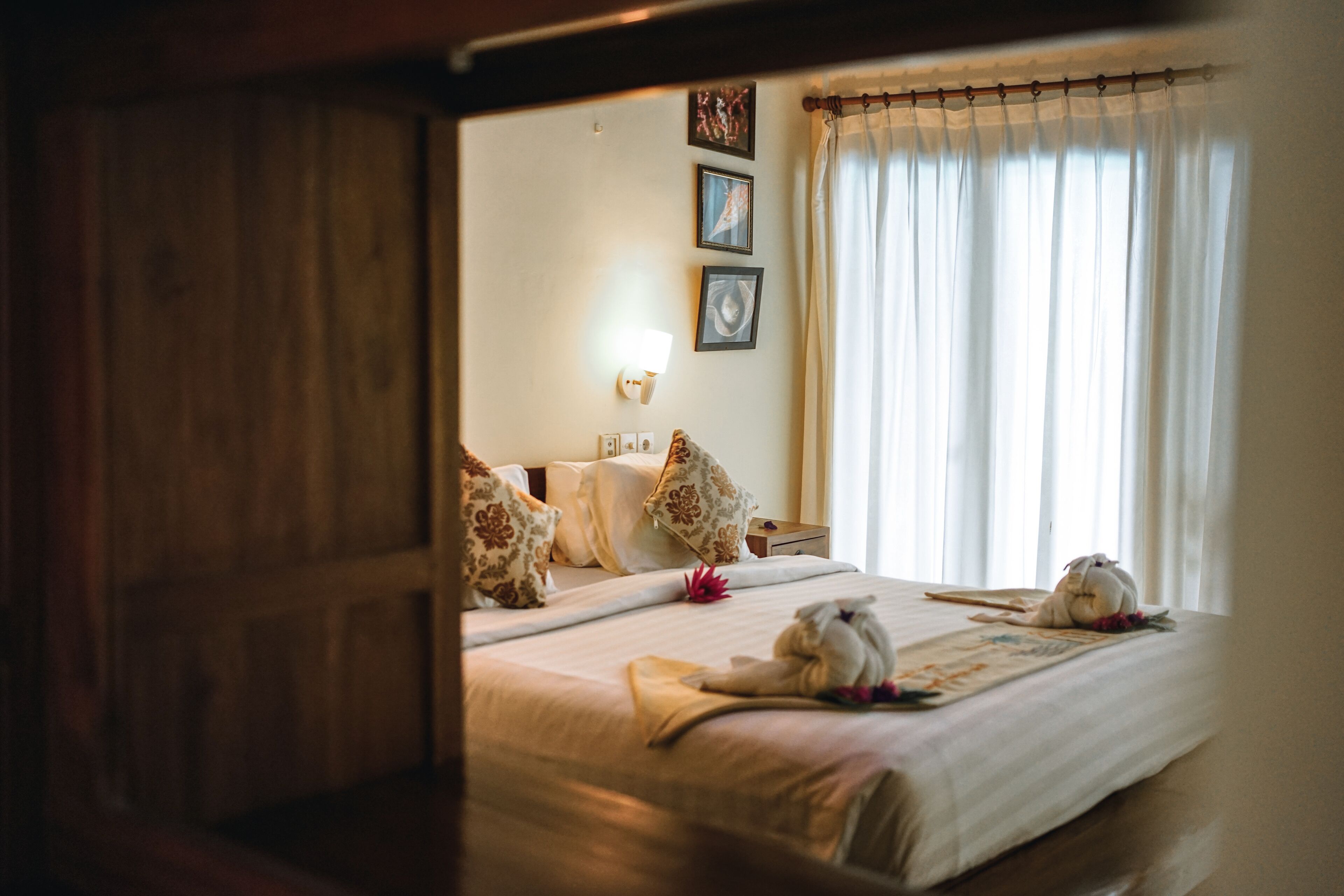 A hotel room with a wooden door, a bed with white linens, decorative pillows, a wall-mounted light, and a window with curtains.