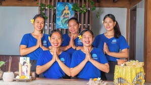 A group of people in blue shirts standing in front of a wooden structure with a painting and a table with a yellow cloth. - Amed Cafe & Hotel Kebun Wayan (Karangasem)