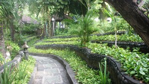 A stone pathway winding through a lush garden with various plants and a traditional structure in the background. - Amed Cafe & Hotel Kebun Wayan (Karangasem)