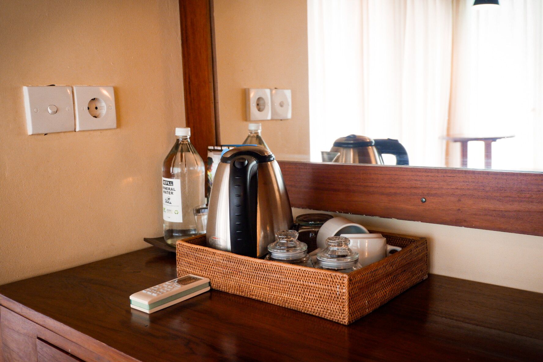 A hotel room with a wooden dresser, a mirror, a bottle of mineral water, a kettle, and a basket containing glasses and a remote control.