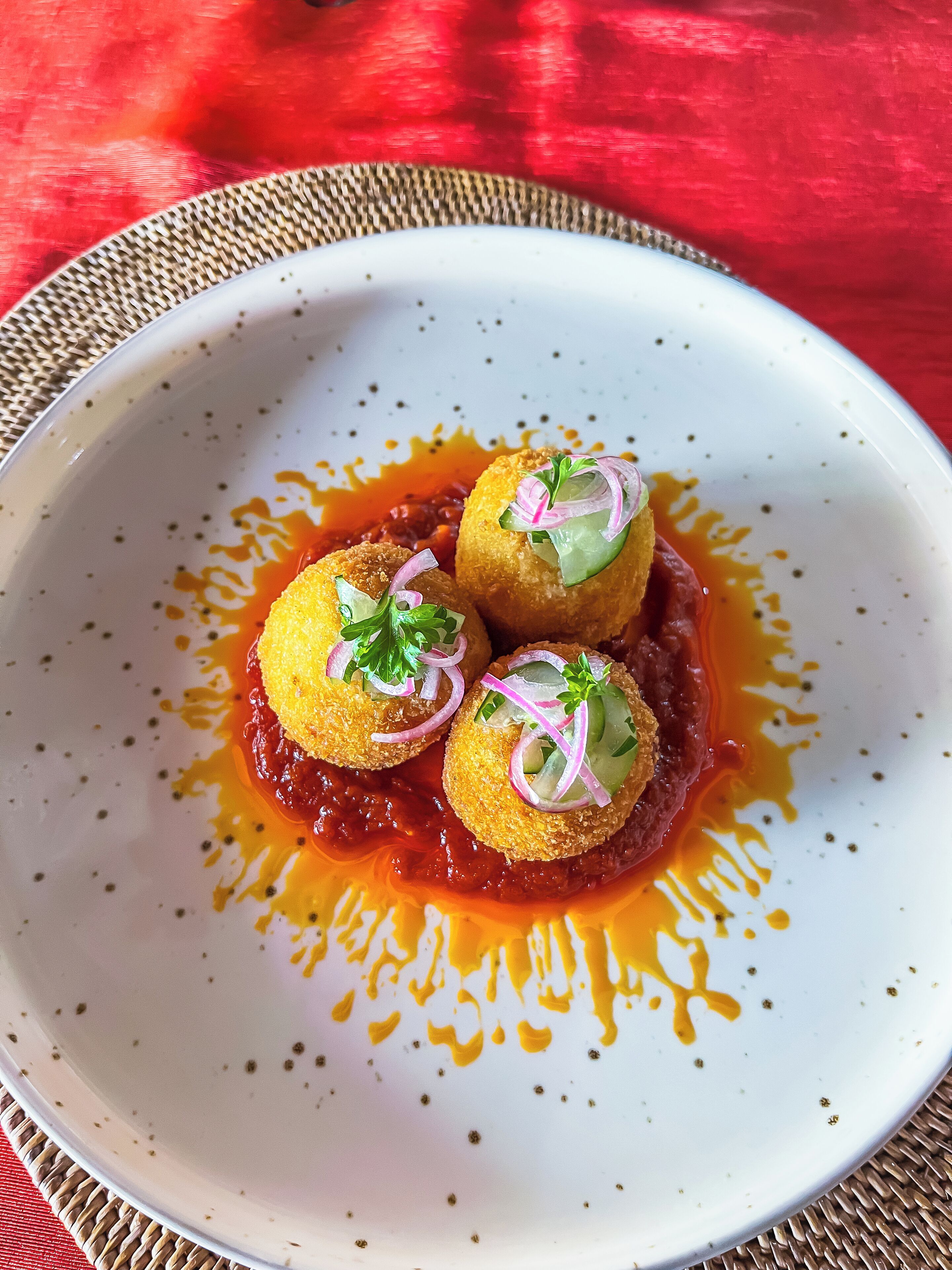 Une assiette contenant trois portions rondes, panées, garnies d’oignons rouges et de verdure, le tout nappé d’une sauce rouge.