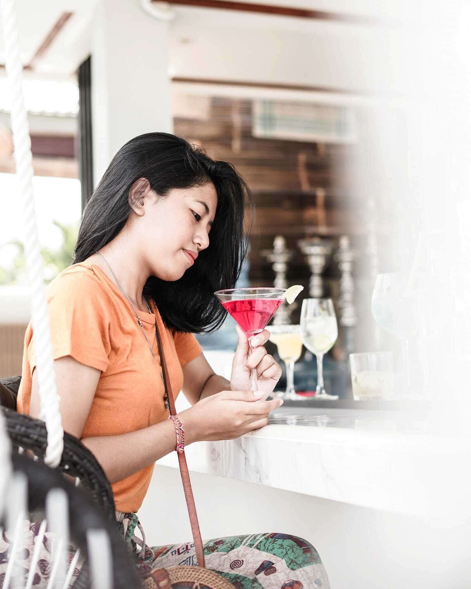 Una persona sosteniendo una copa de cóctel en un bar, con una copa de vino y una botella al fondo.