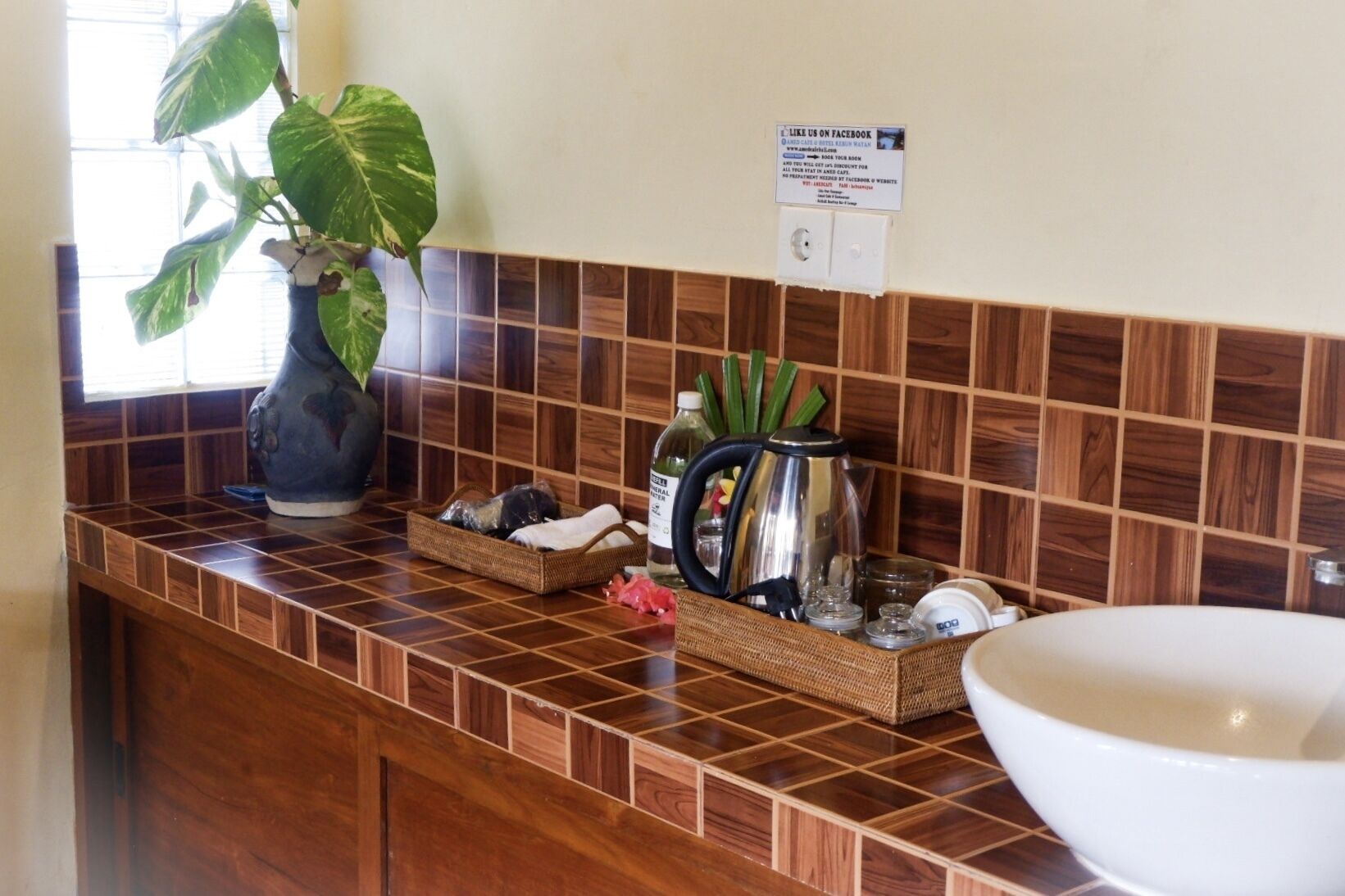 Un baño con tocador de madera, un lavamanos blanco grande, un florero con hojas verdes y un cartel que promociona Facebook.