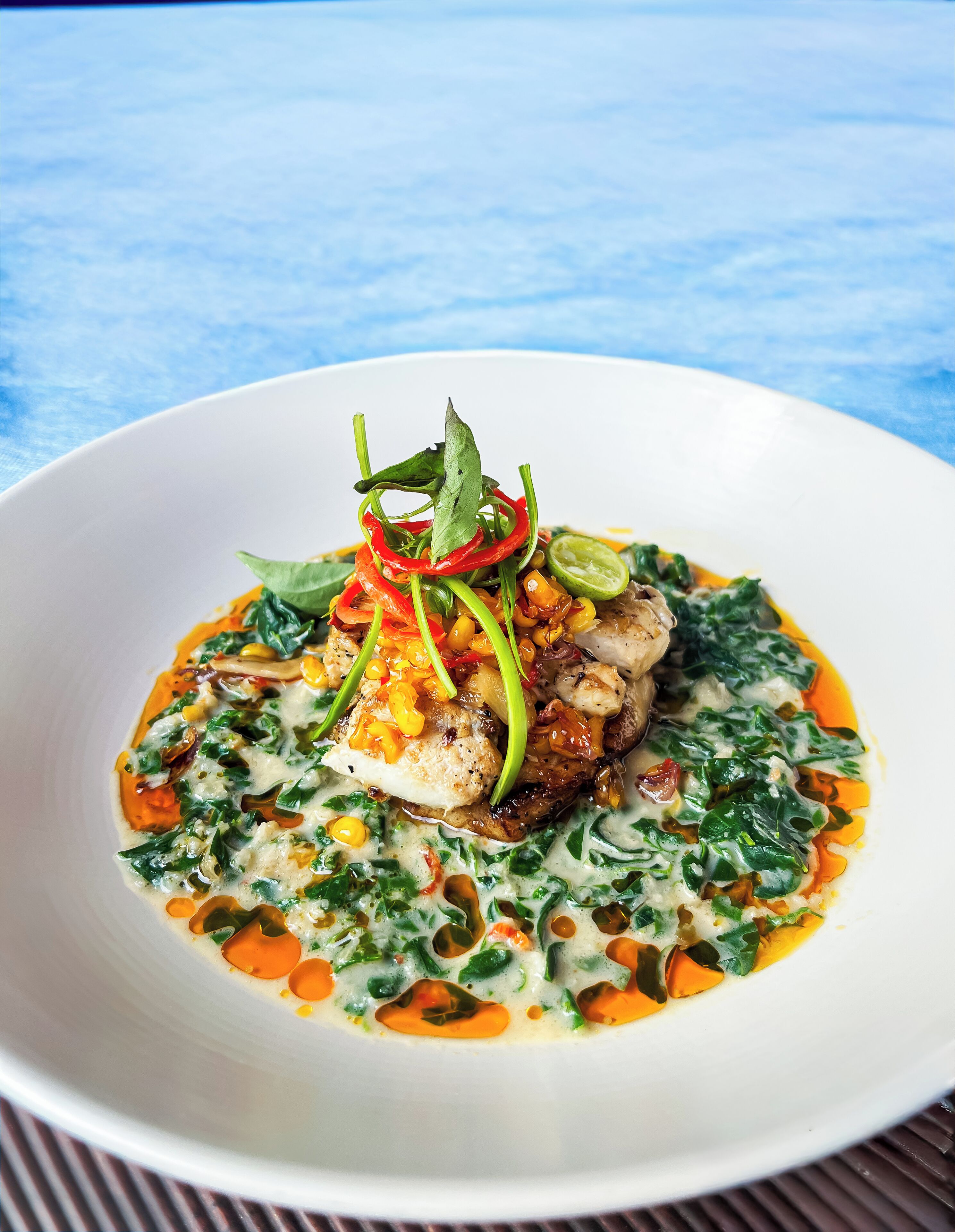 Une assiette blanche contenant un plat de poisson, agrémenté de piments rouges et d’herbes vertes, le tout sur un fond bleu.