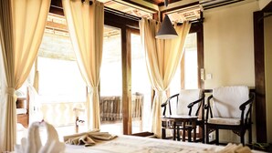 A room with a bed, two chairs, a table, and a balcony with curtains. - Amed Cafe & Hotel Kebun Wayan (Karangasem)