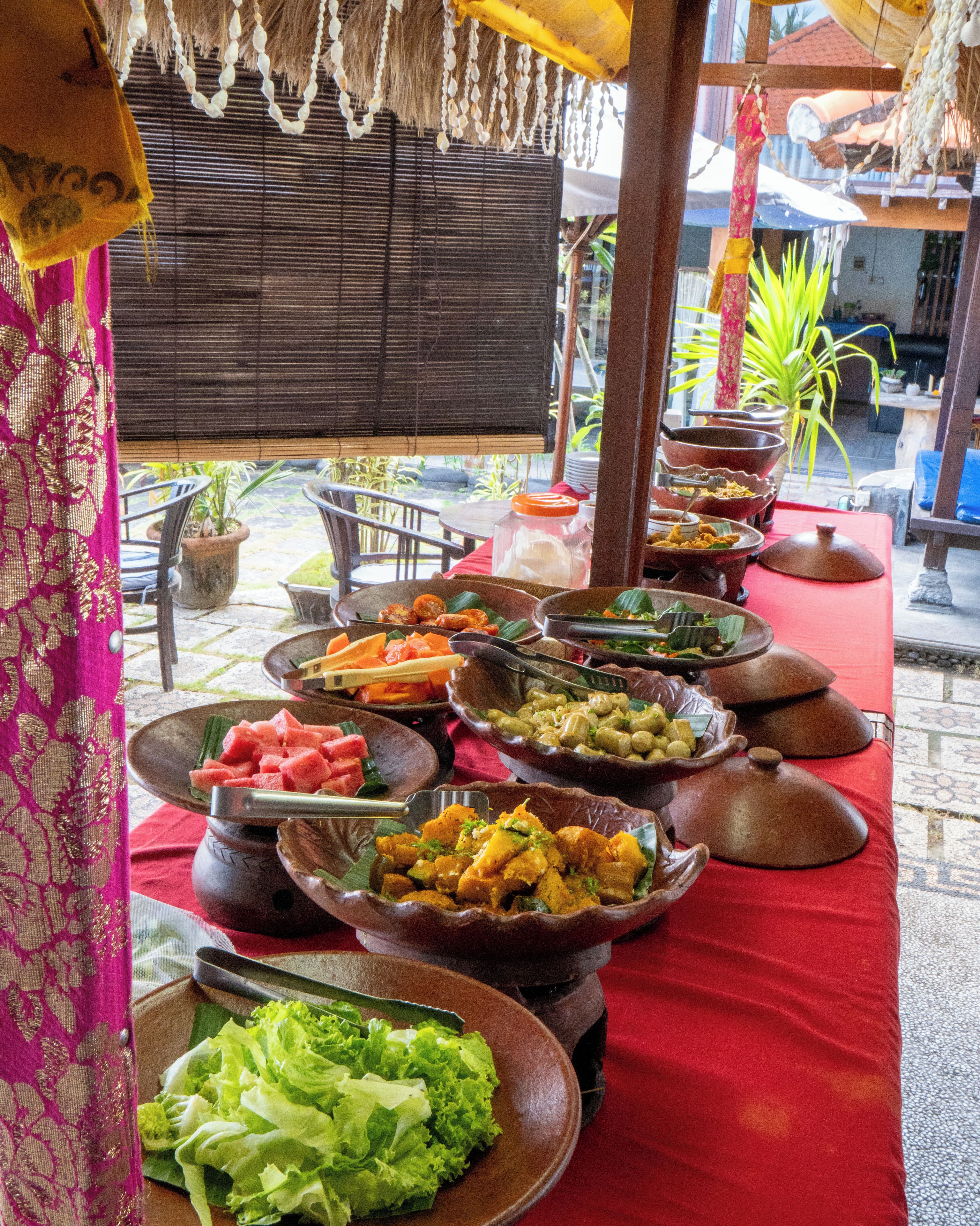 Une table de buffet garnie de différents plats et de saladiers, une nappe rouge et une structure surmontée d’un toit de chaume.