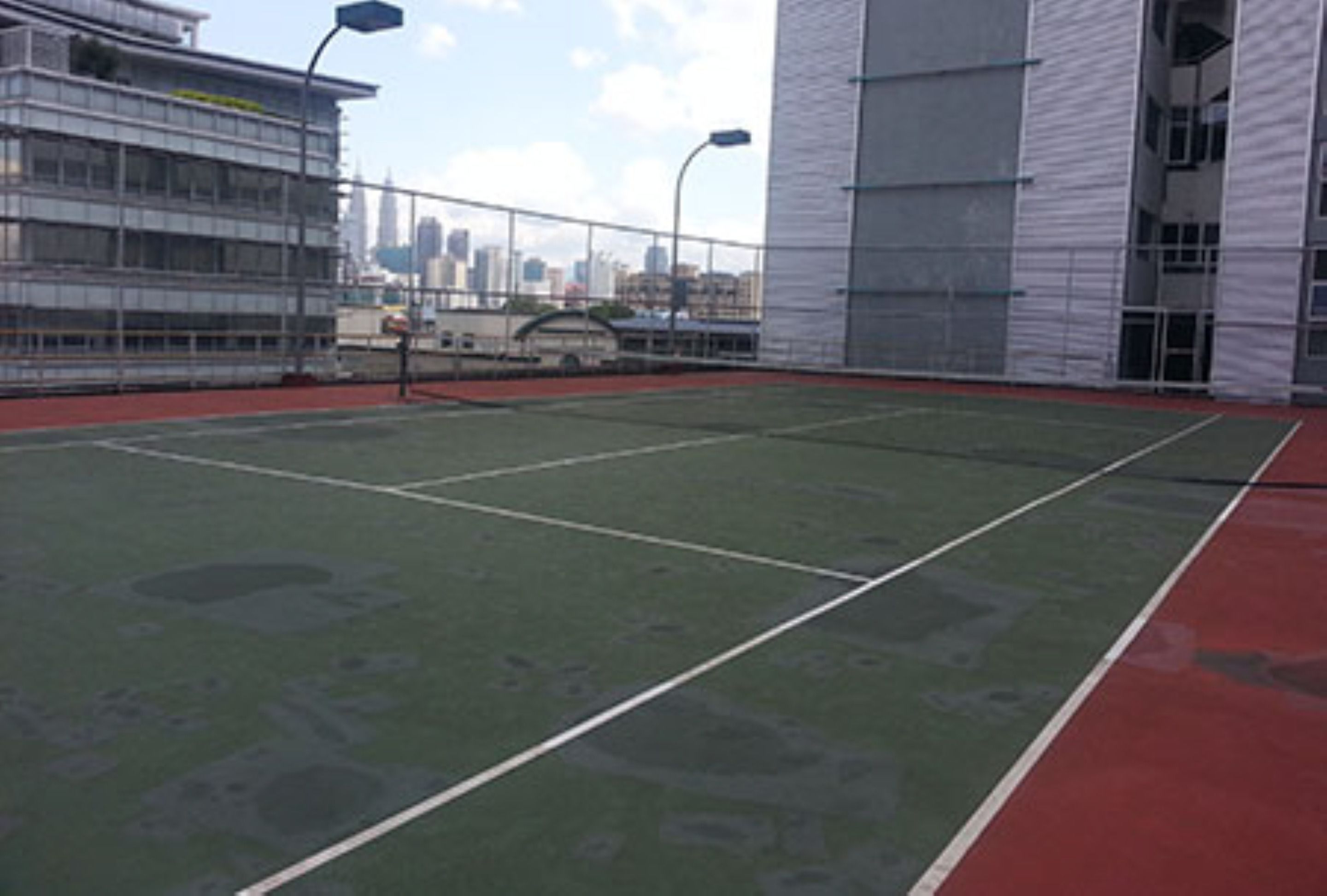 An outdoor tennis court with a red and green surface, surrounded by a metal fence and a building with a white facade.
