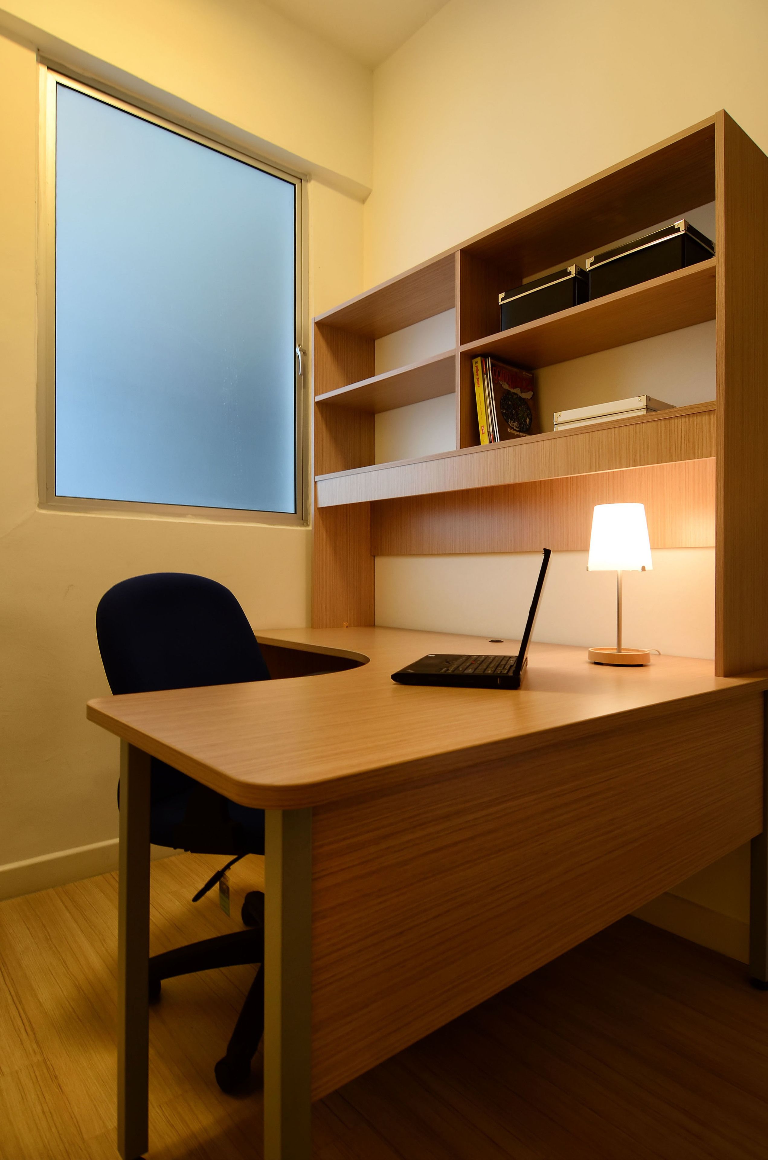 Un escritorio con una computadora portátil y una lámpara, una silla, y una estantería con libros y una caja.