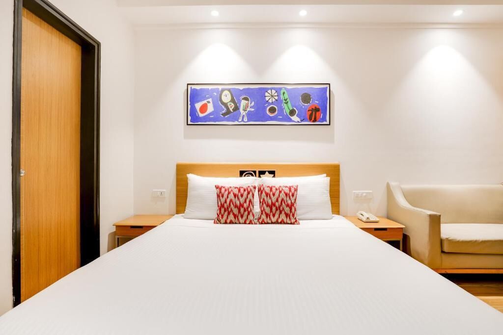 Habitación de hotel con una cama grande, cabecera de madera, mesita de noche con teléfono, una silla y una obra de arte en la pared sobre la cama.