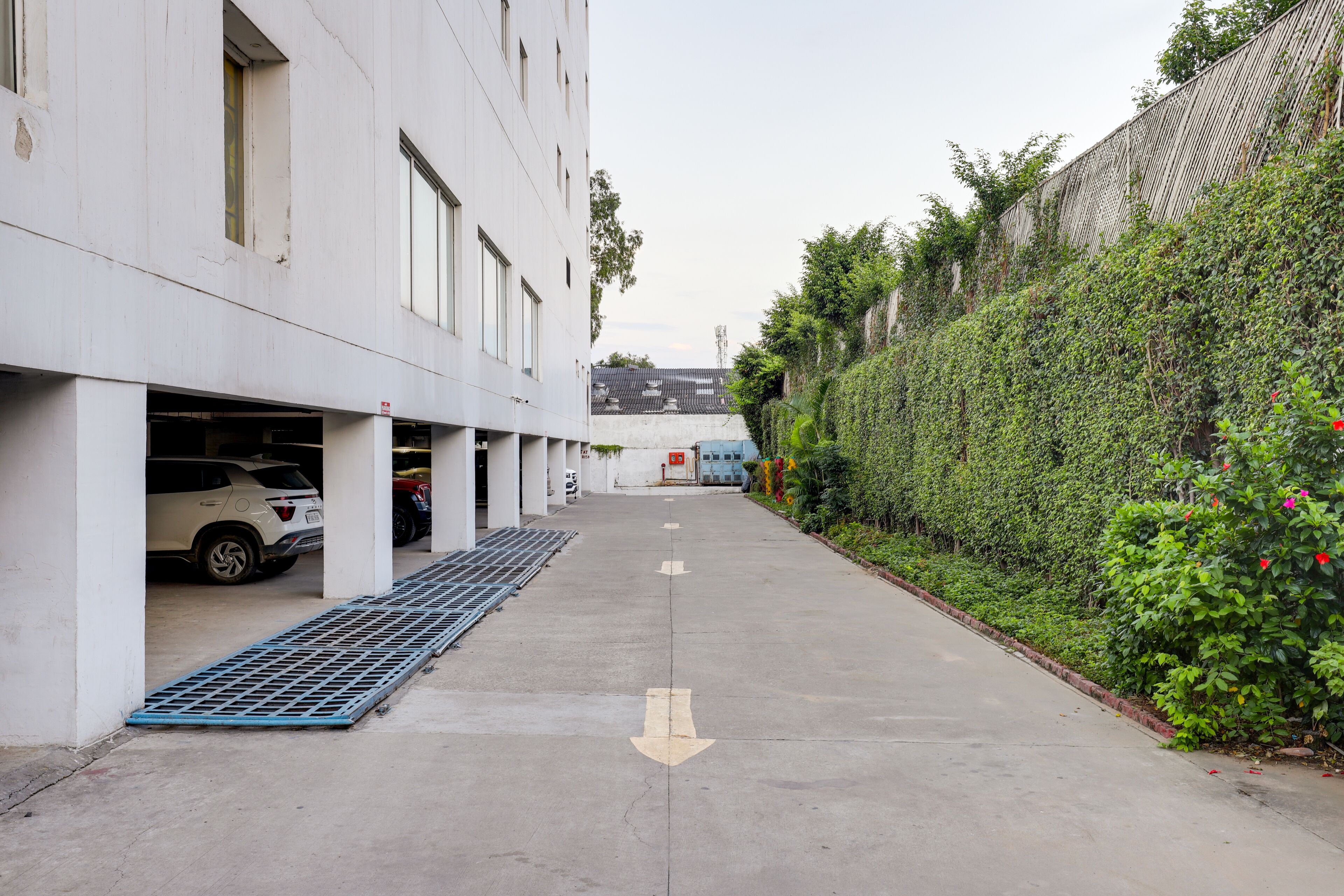 Un garaje para estacionar con un edificio blanco, una rejilla de drenaje azul y un seto verde.