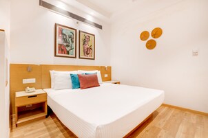 A hotel room with a large bed, wooden headboard, two bedside tables, a nightstand, and a wall-mounted air conditioning unit.