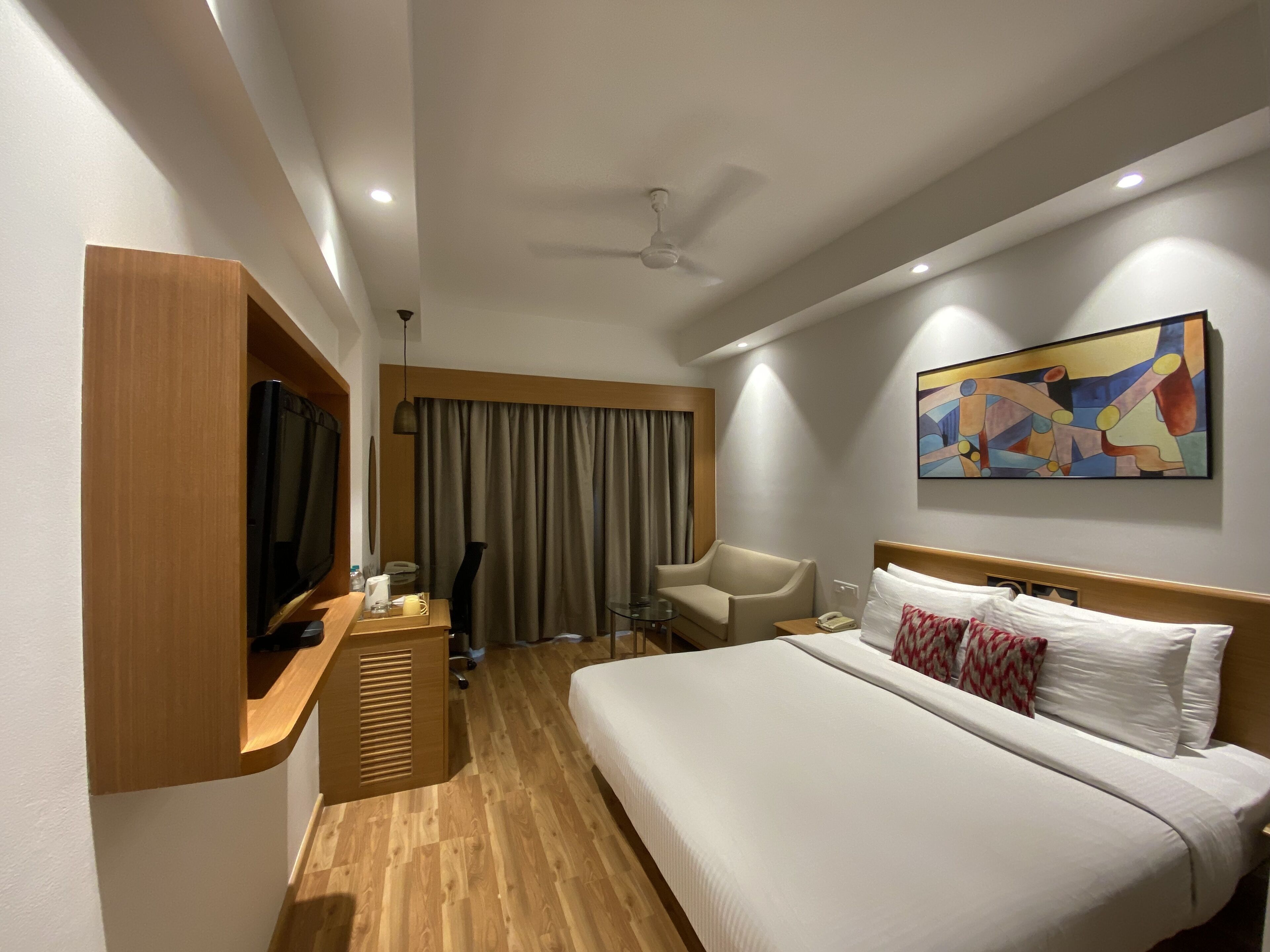 Habitación de hotel con cama, una silla, escritorio con televisor, ventilador de techo y un cuadro en la pared.