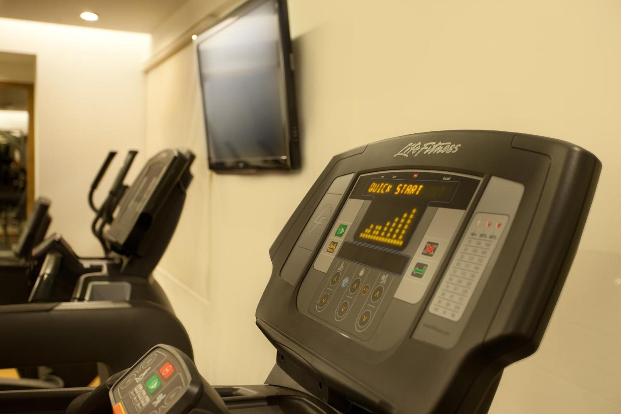 A treadmill with a digital display and control panel, located in a room with a television on the wall.