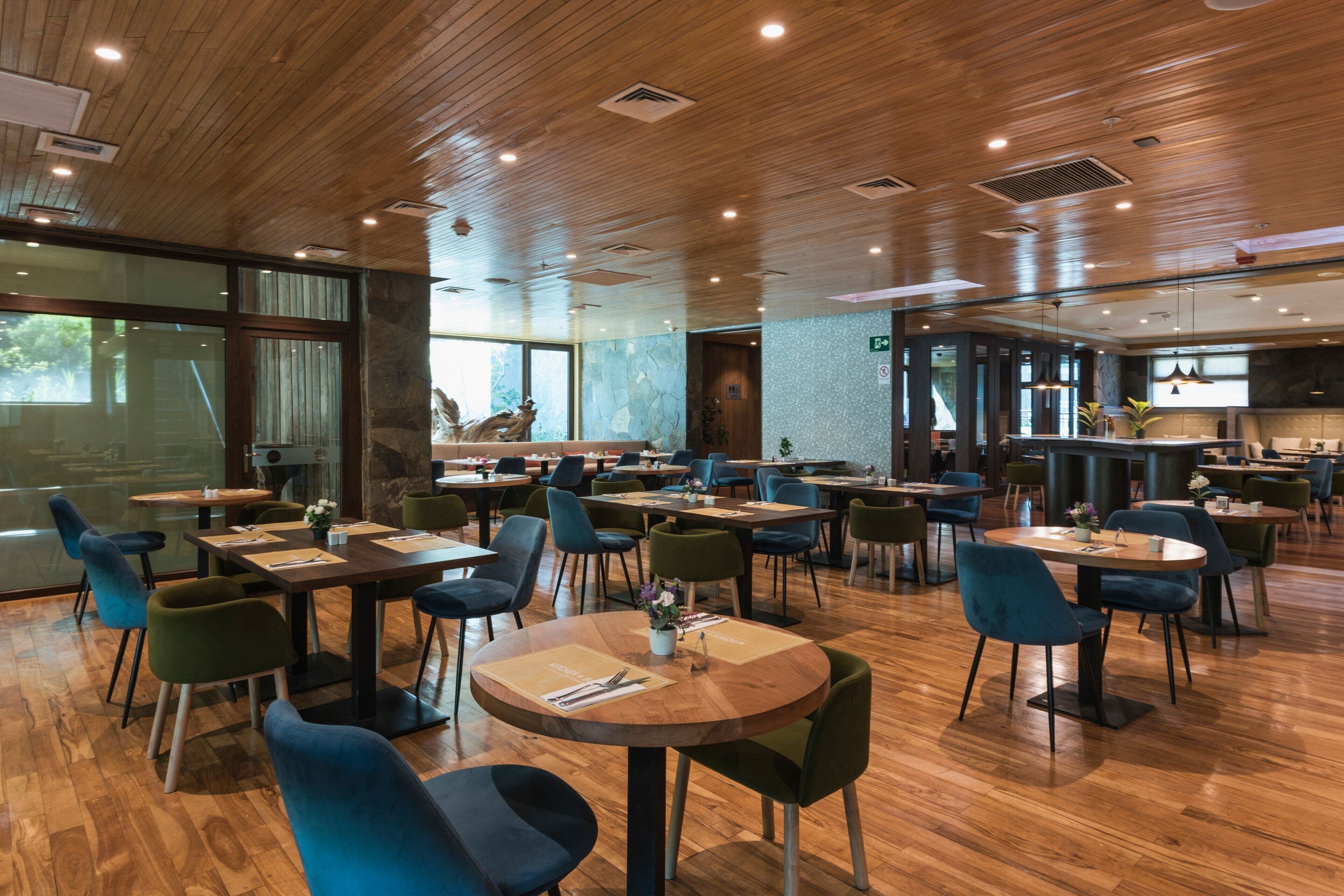 A spacious restaurant with wooden flooring, green and blue chairs, and a modern interior design.