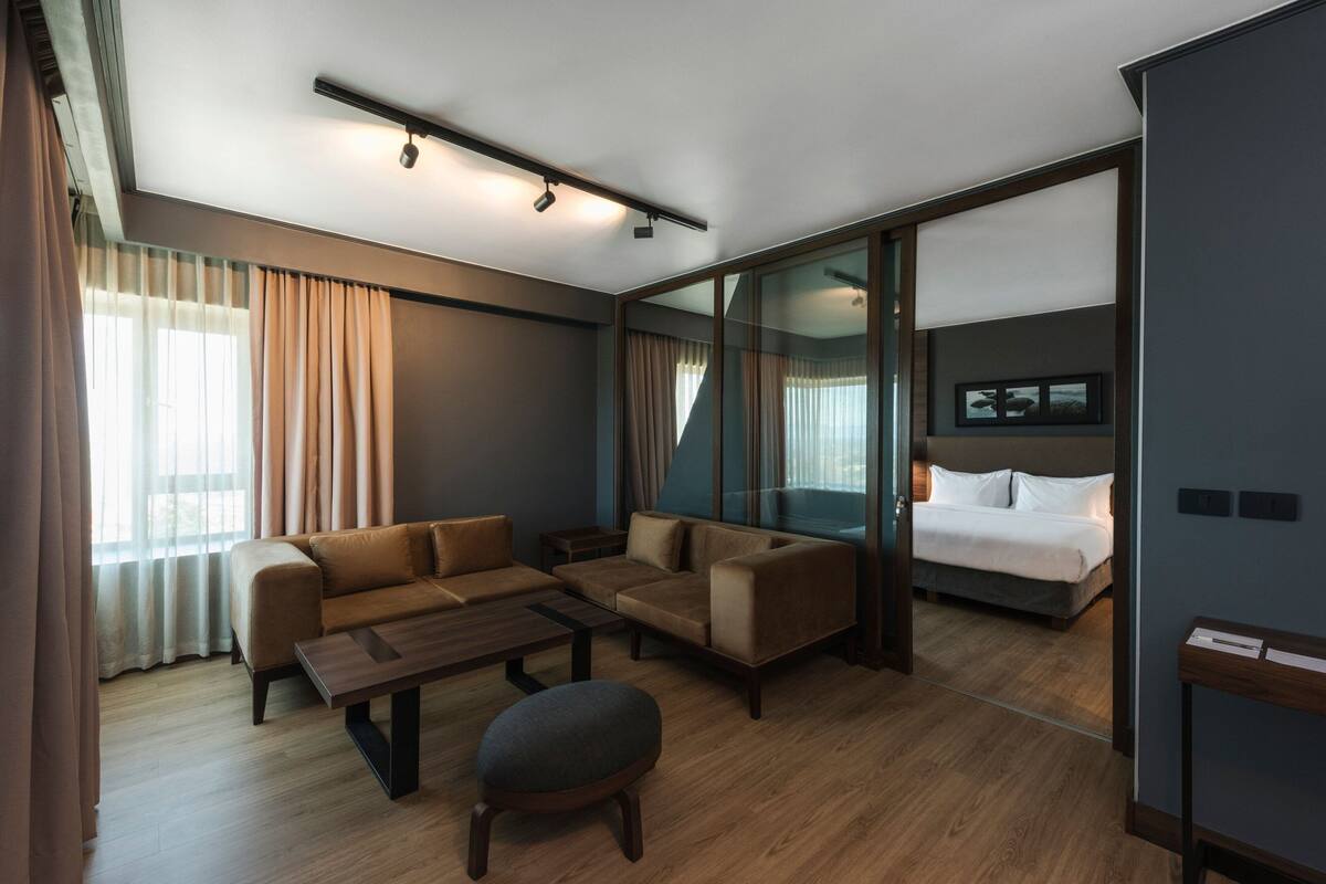Ein modernes Hotelzimmer mit einer Sofagarnitur, einem Bett, einem Holztisch und einer Glastrennwand.