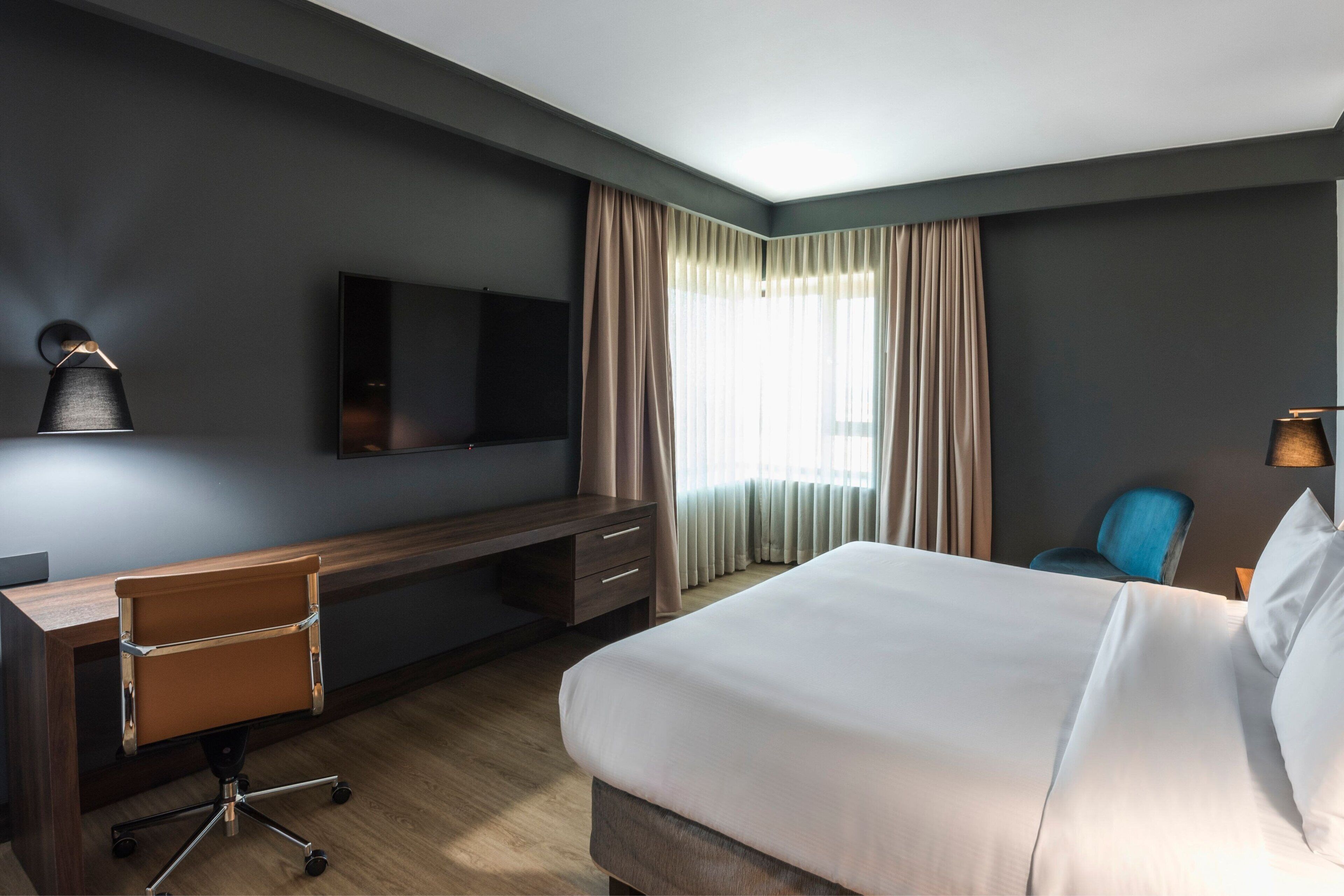 A hotel room with a large bed, a desk with a chair, a flat-screen TV, and a window with curtains.