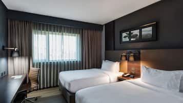 Quarto de hotel com duas camas, uma mesa, uma cadeira, um telefone e uma janela com cortinas.