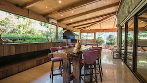Un amplio espacio exterior con comedor, una mesa y sillas de madera rústica, rodeado de grandes ventanales con vistas a zonas verdes.