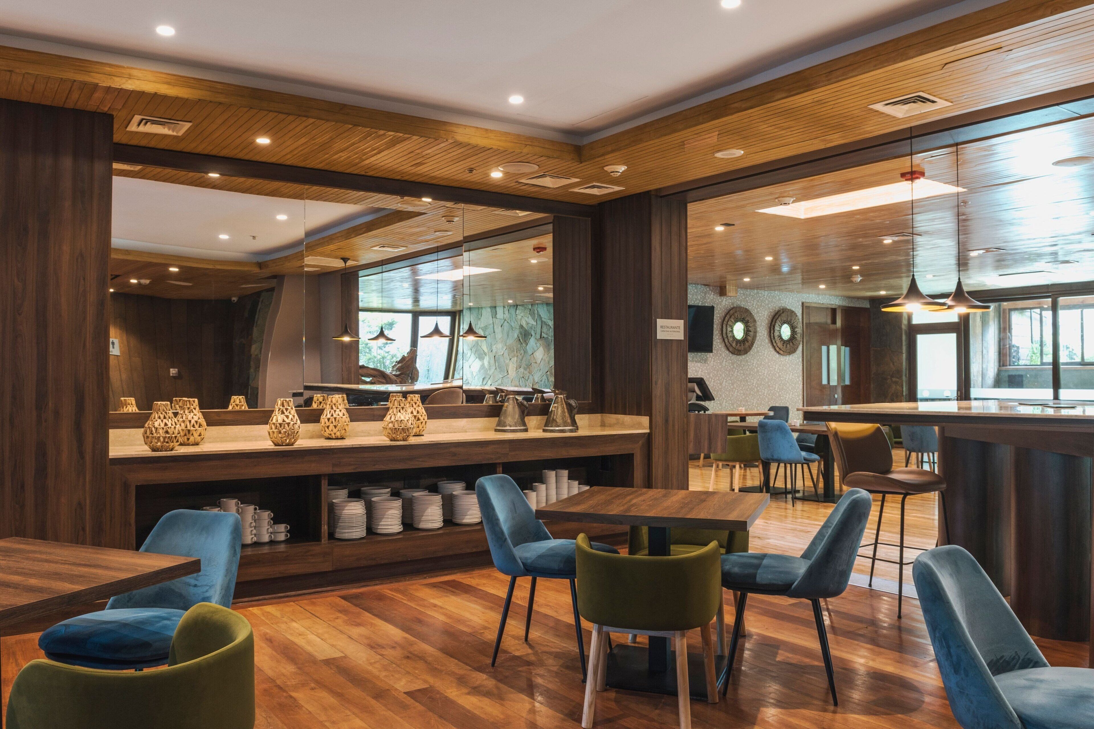 A modern restaurant interior with wooden flooring, blue and green chairs, a bar area, and a large window providing natural light.