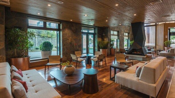 Un hallo amplio con pared de piedra, techo de madera y ventanas grandes. Hay varias zonas de estar con sofás y sillones.