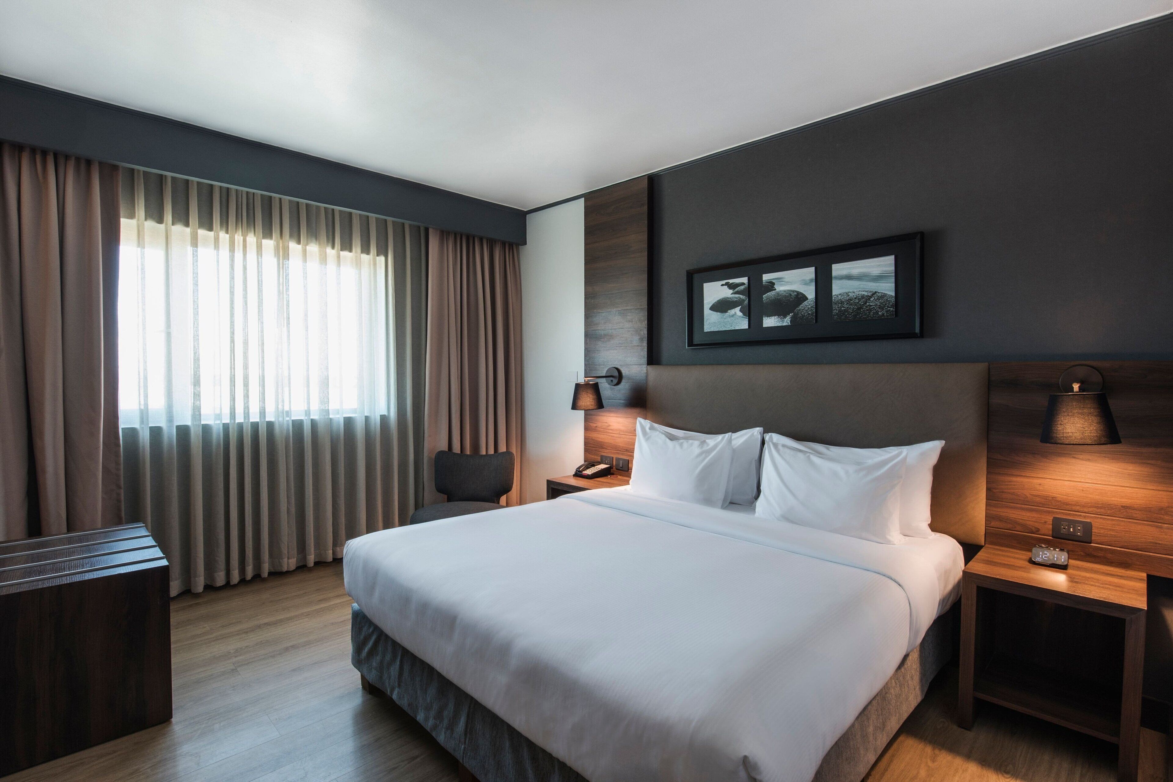 Habitación de hotel moderna con una cama grande, una mesita de noche con lámpara, una mesita auxiliar y una silla.