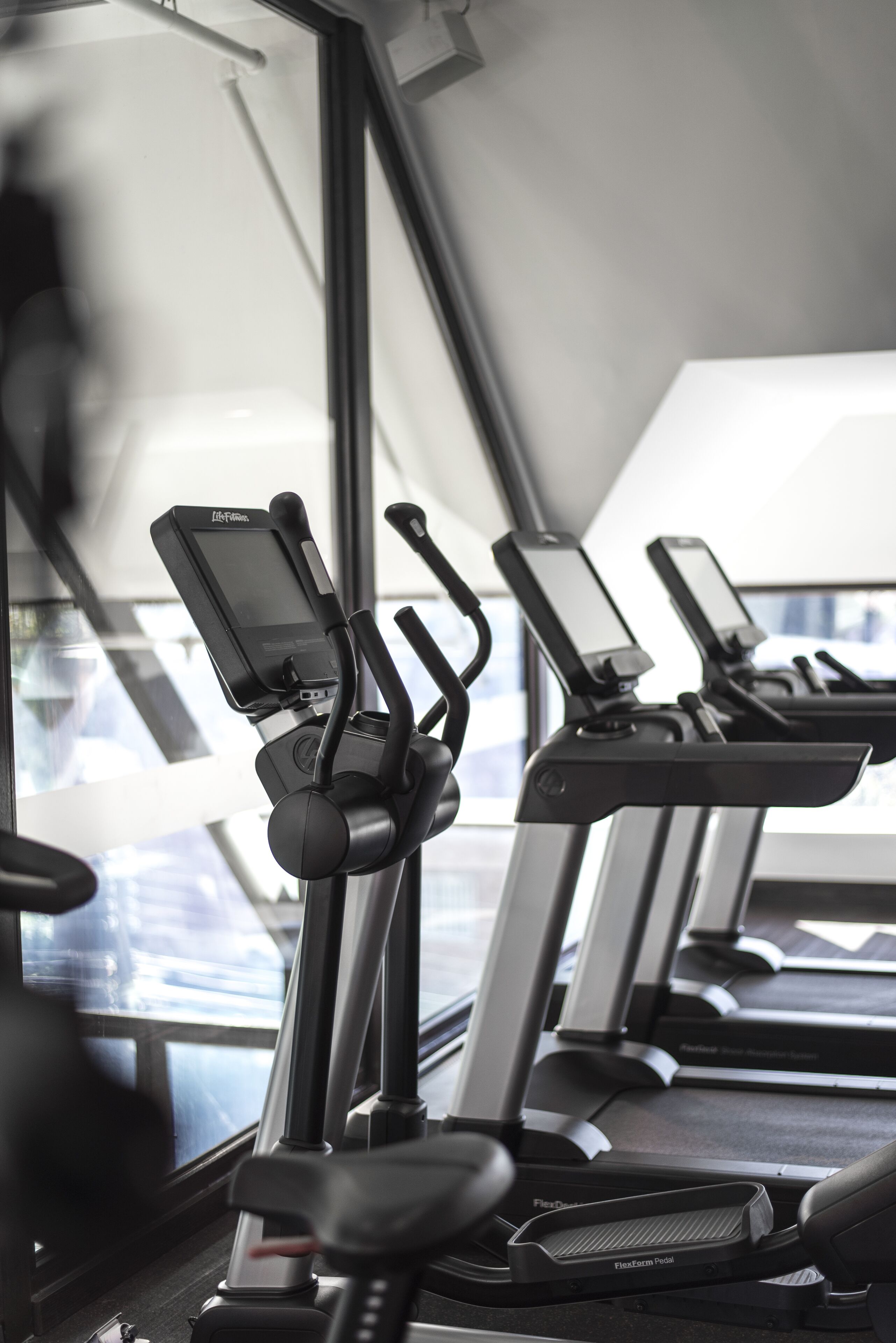 A row of stationary bikes with digital screens, positioned in front of a large window.