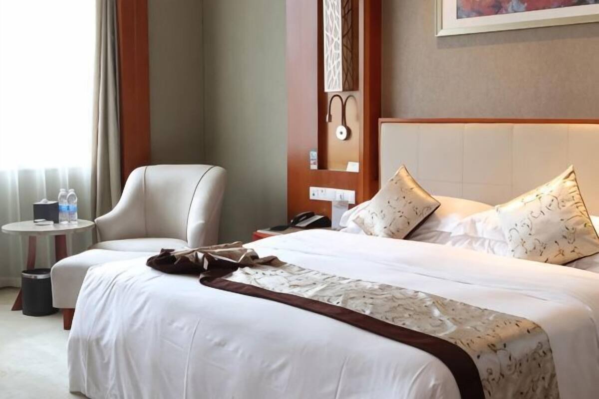 Une chambre d’hôtel avec un lit, une chaise, une petite table avec un téléphone et une bouteille d’eau, et une horloge fixée au mur.