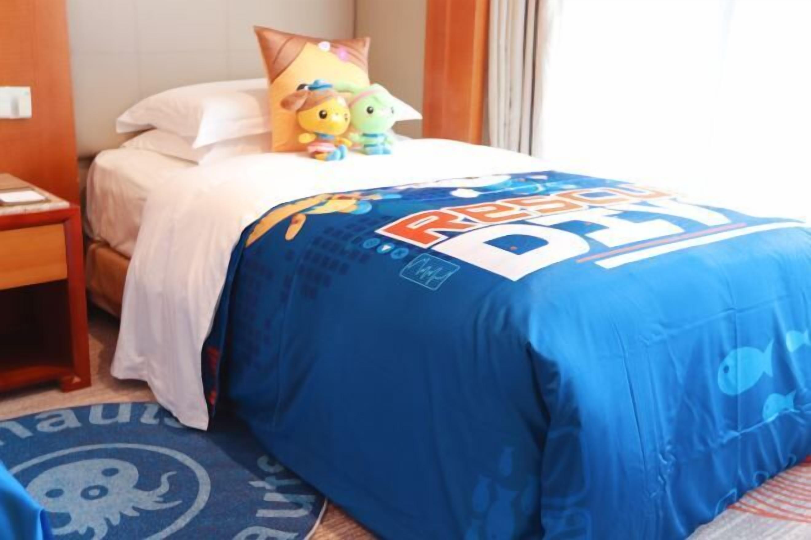 A hotel room with a bed, a nightstand, a blue bedspread with a cartoon character, and a small rug.