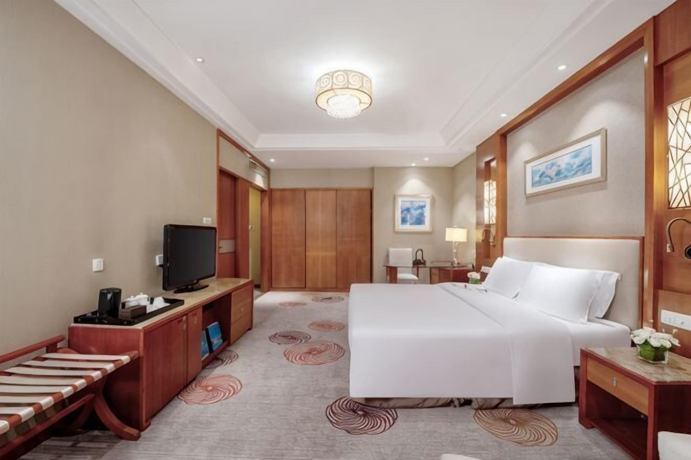 A hotel room with a bed, a desk with a television, a desk lamp, a nightstand, and a wall-mounted artwork.