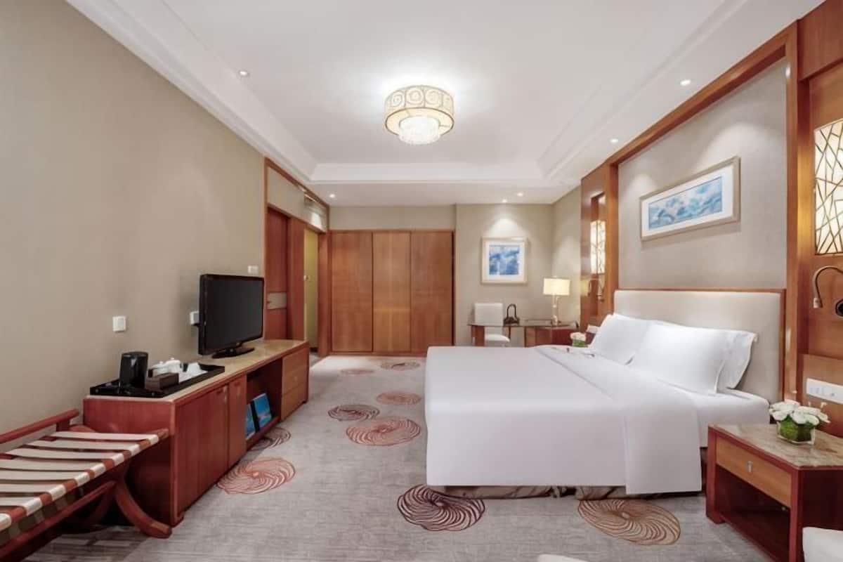 A hotel room with a bed, a desk with a television, a desk lamp, a nightstand, and a wall-mounted artwork.
