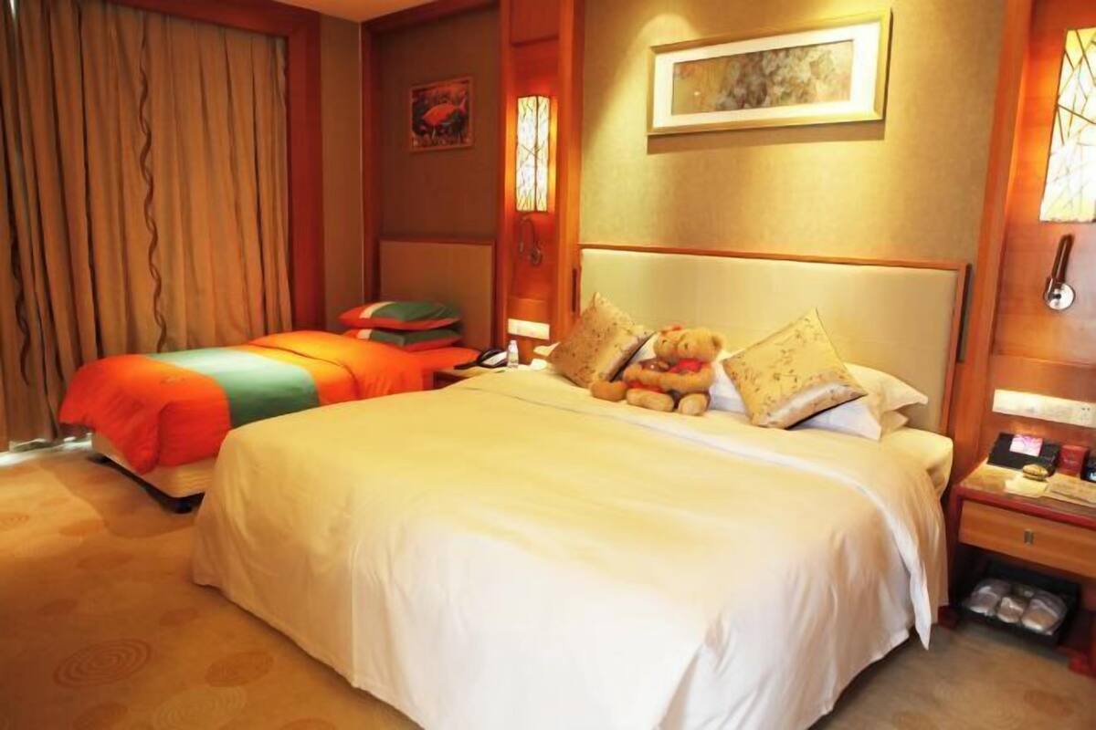Une chambre d’hôtel avec un grand lit, un lit plus petit, une table de chevet, une lampe et une fenêtre avec des rideaux.