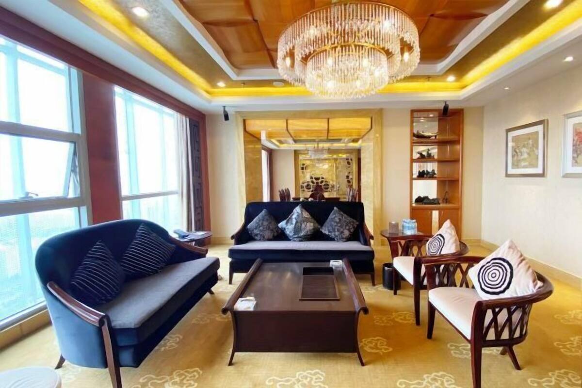 Une chambre d’hôtel équipée d’un canapé, de fauteuils, d’une table basse, d’un lustre et d’un présentoir à chaussures.