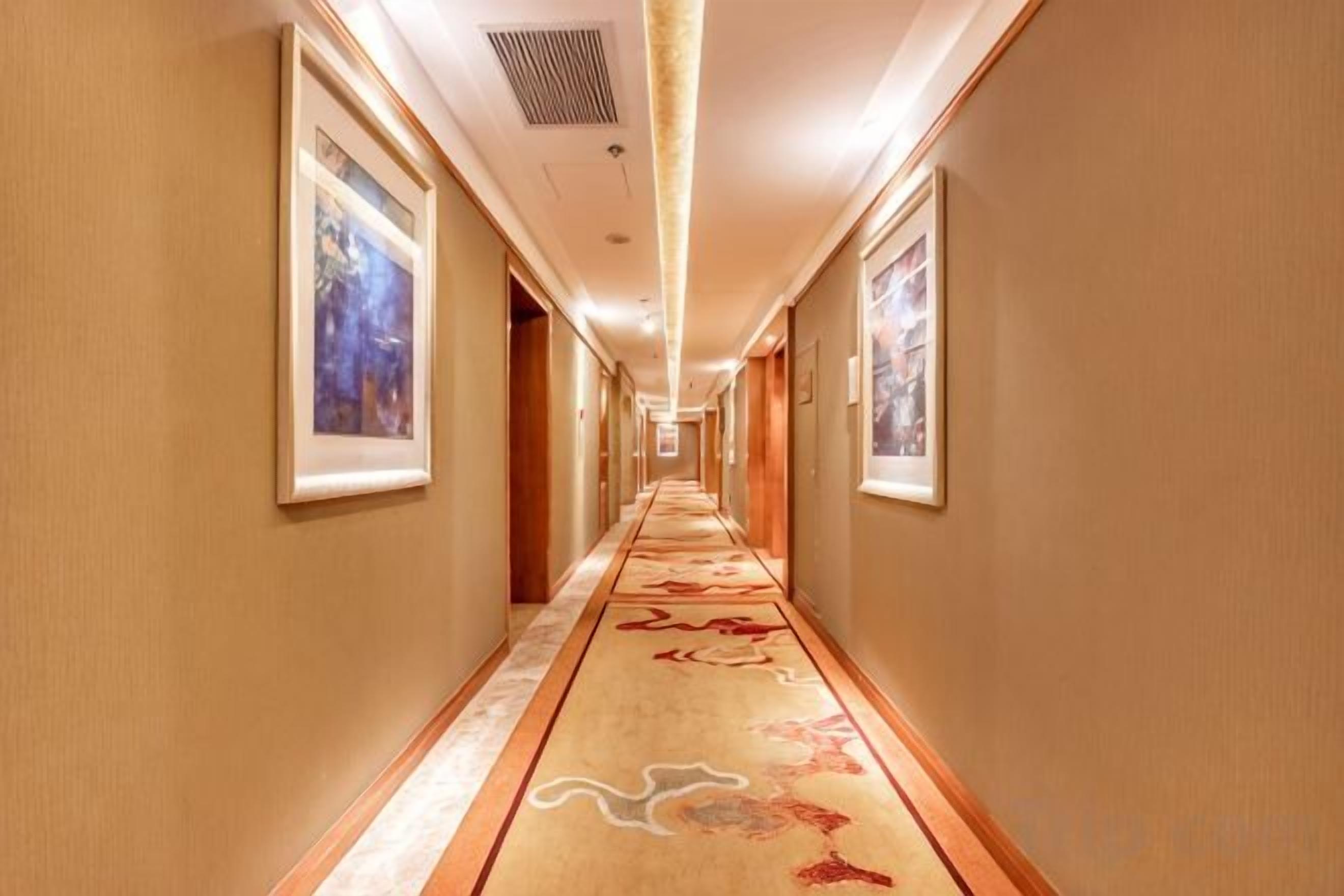 A hotel hallway with framed artwork on the walls, carpeted flooring, and a long corridor leading to a door.