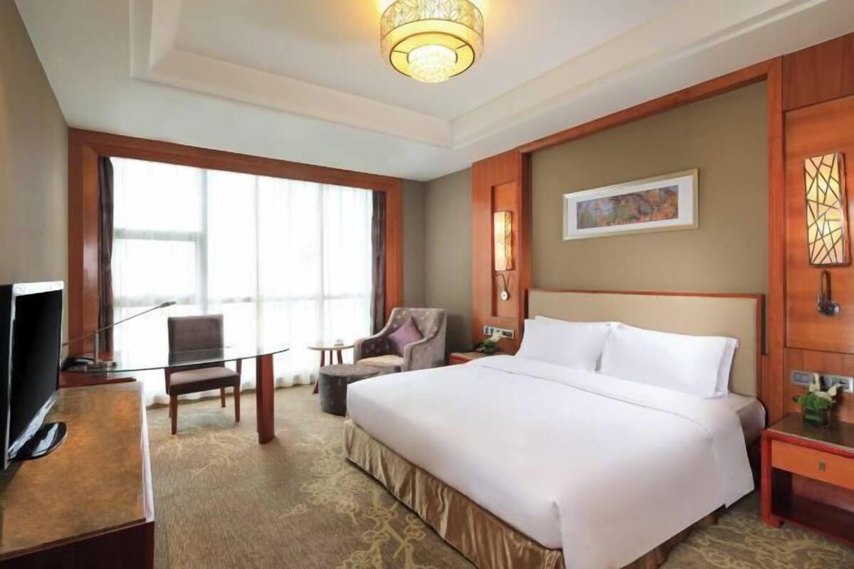 Une chambre d’hôtel avec un grand lit, un bureau avec une télévision, une chaise, une petite table et une fenêtre avec des rideaux.