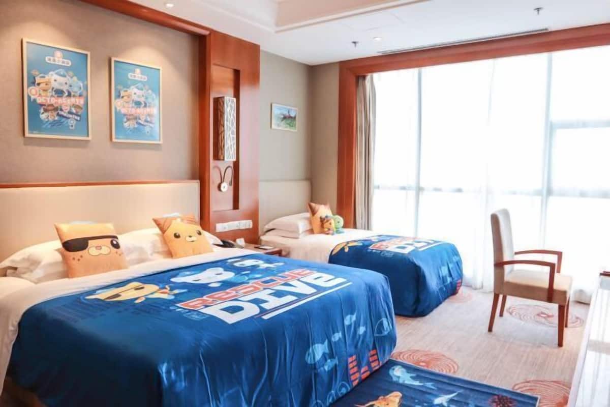 Une chambre d’hôtel avec deux lits, chacun recouvert d’une courtepointe bleue sur le thème de la plongée, dotée d’une tête de lit en bois, d’une grande fenêtre avec des rideaux et d’une chaise en bois.