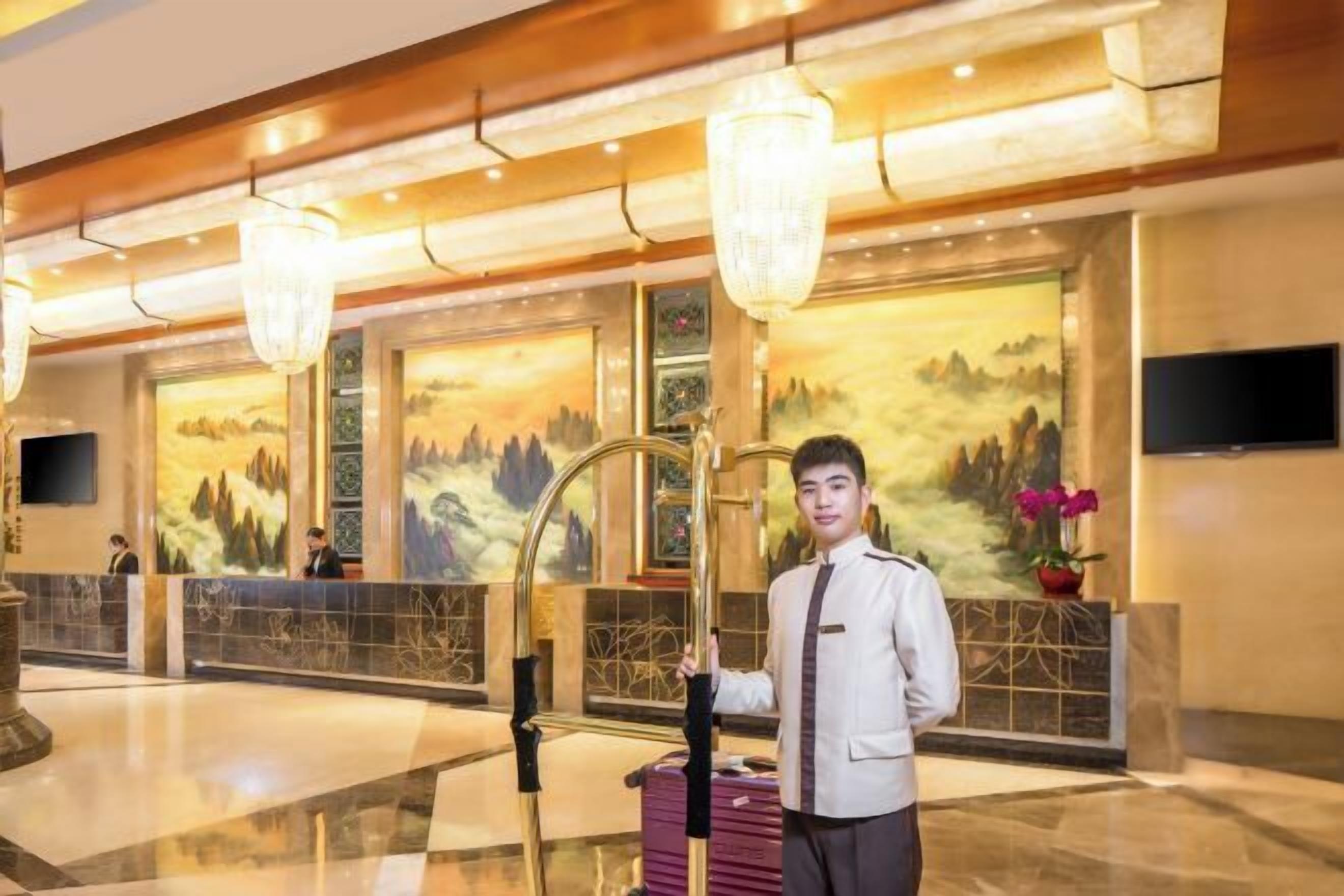A hotel lobby with a reception desk, a large chandelier, and a painting of a mountainous landscape on the wall.