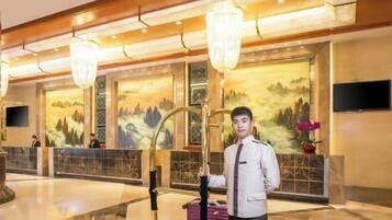 A hotel lobby with a reception desk, a large chandelier, and a painting of a mountainous landscape on the wall.