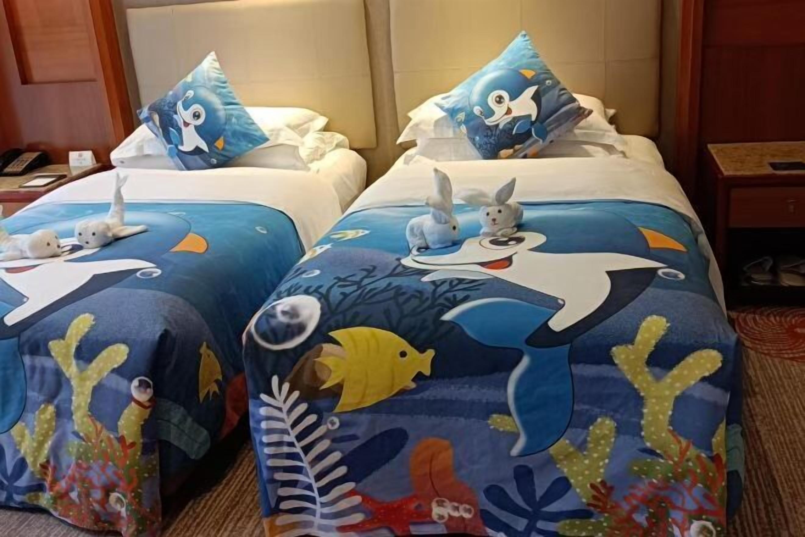 A hotel room with two beds, each adorned with a blue and white ocean-themed bedspread featuring sea creatures and a pillow with a blue bird design.