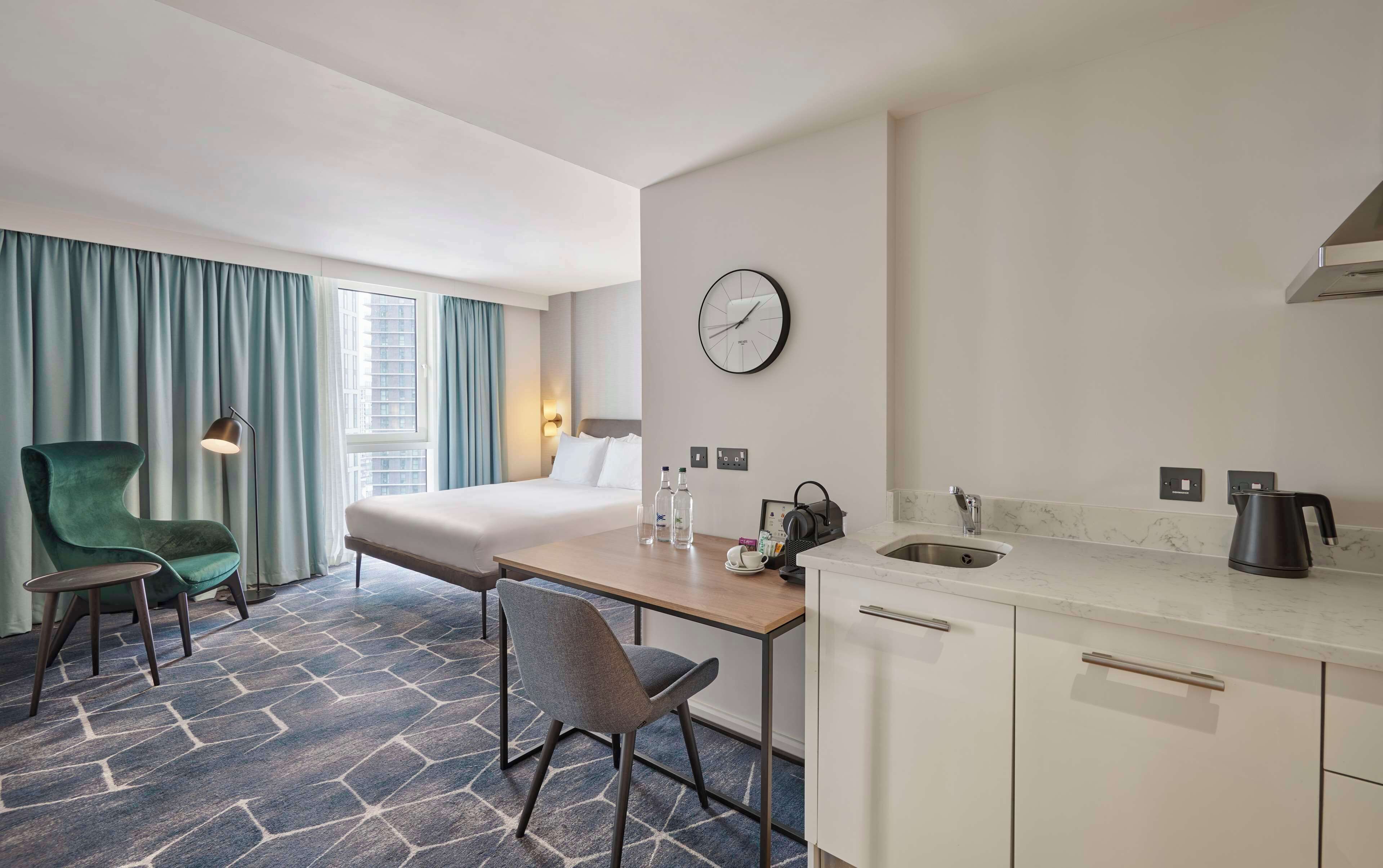 Habitación de hotel con cama, escritorio, silla, reloj, lavamanos y ventana con cortinas.