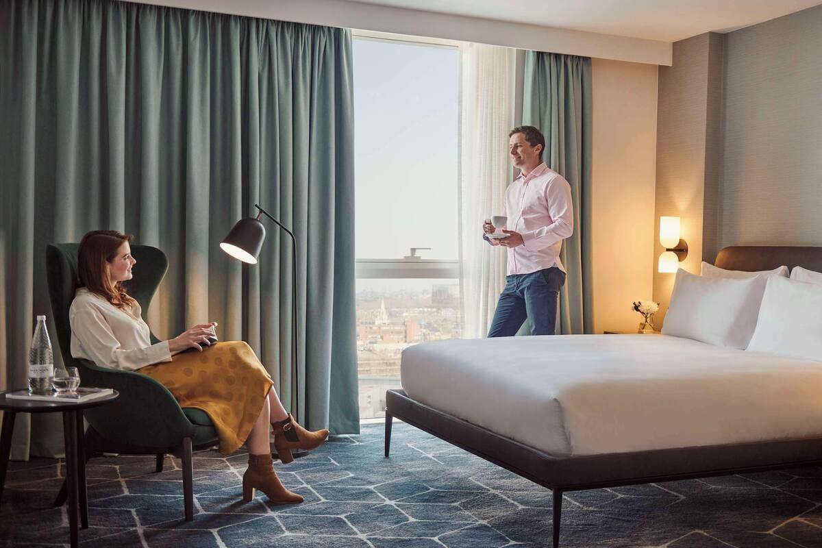 Ein Hotelzimmer mit Bett, Stuhl, Tisch, einer Flasche und einem Glas sowie Blick auf die Stadt durch große Fenster.