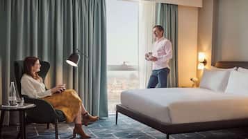 A hotel room with a bed, a chair, a table with a bottle and glass, and a view of the city through large windows.