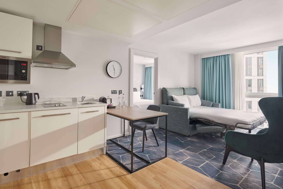 Ein kompaktes Hotelzimmer mit Küchenzeile, einem Bett, einer Sitzecke und Blick auf die Stadt.