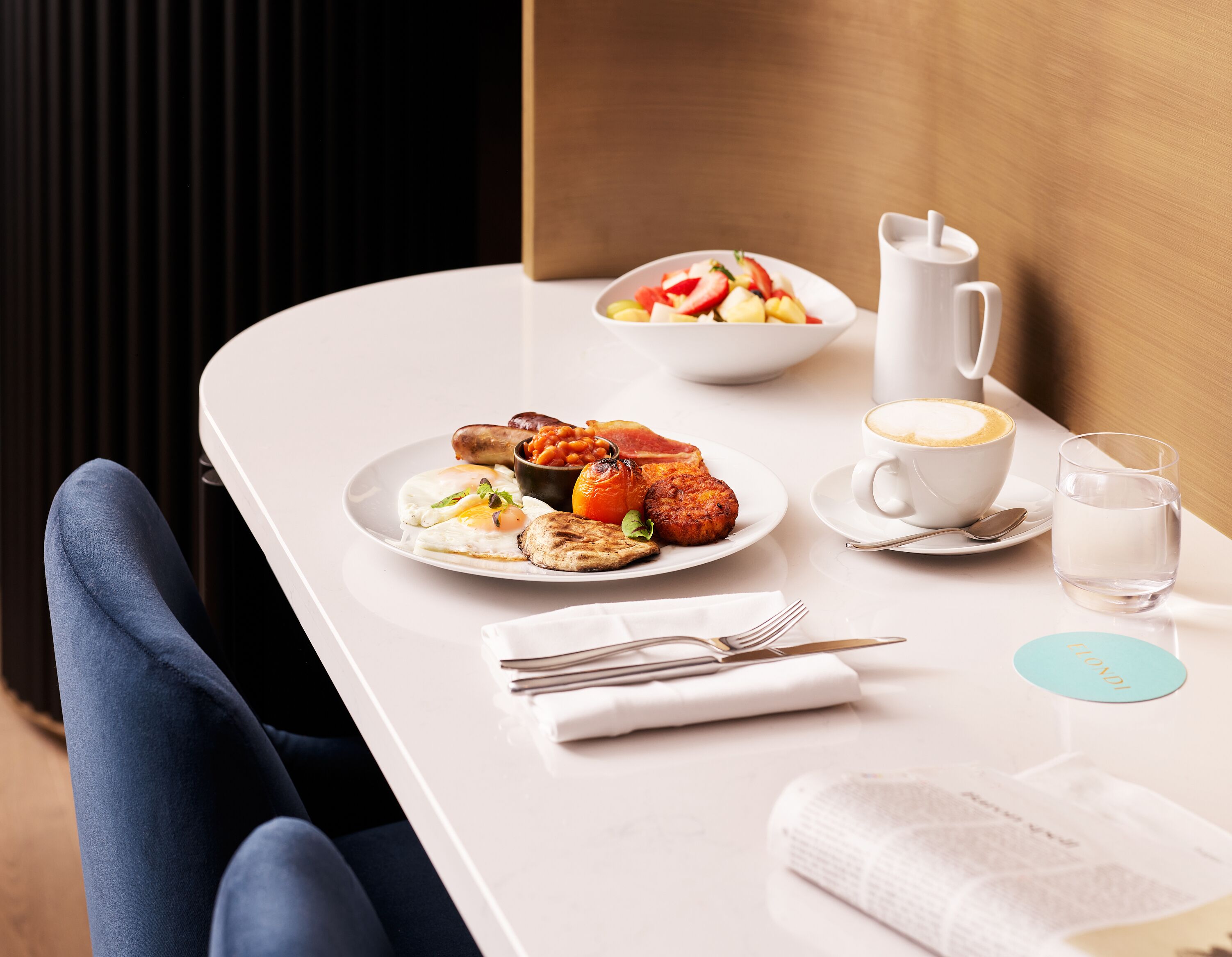 Un desayuno con huevos, salchichas y frijoles horneados en un plato blanco, una taza de café y un vaso de agua sobre una mesa blanca.