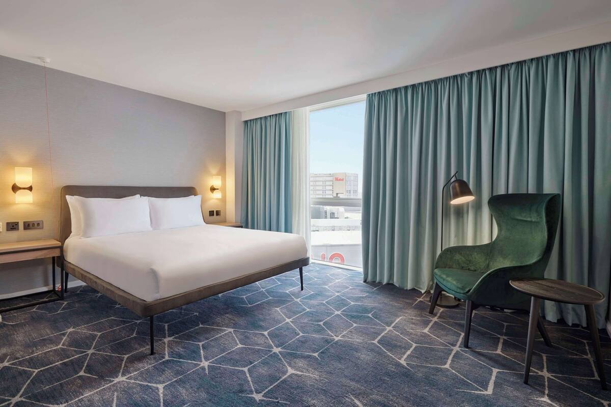 Ein Hotelzimmer mit einem großen Bett, einem Sessel, einem Schreibtisch und Blick auf die Stadt durch das Fenster.